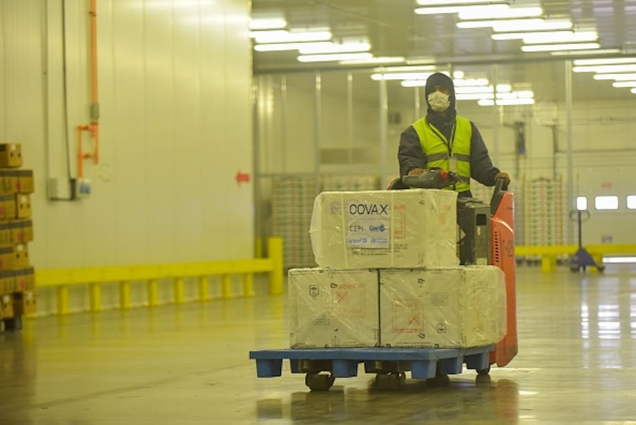 Person wearing protective clothing and face mask sits on a motorised trolley loaded with packages marked COVAX