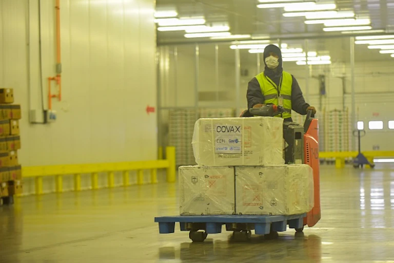 Person wearing protective clothing and face mask sits on a motorised trolley loaded with packages marked COVAX
