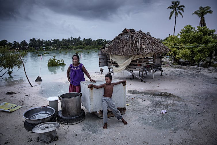 A woman and boy stand outside a home on stilts with cooking pots on the beach where water has risen.