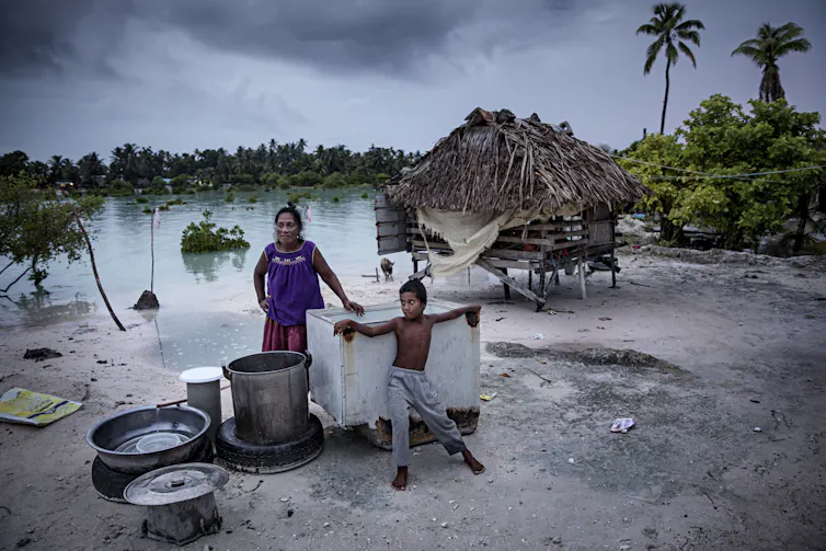 A woman and boy stand outside a home on stilts with cooking pots on the beach where water has risen.