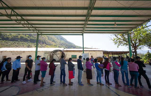 People stand in a line wearing face masks, under a shelter, with mountains in the background