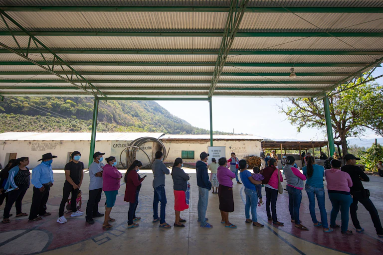 People stand in a line wearing face masks, under a shelter, with mountains in the background