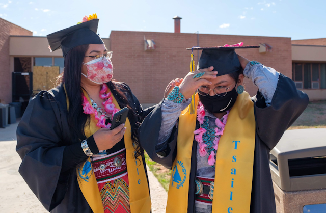Two college graduates wear caps, gowns, masks and Native American accessories