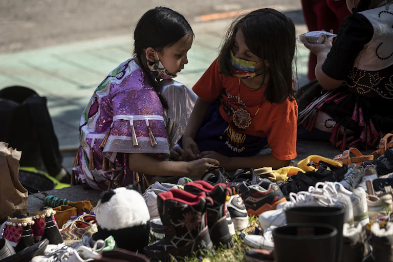 Two children sit in front of a collection of shoes