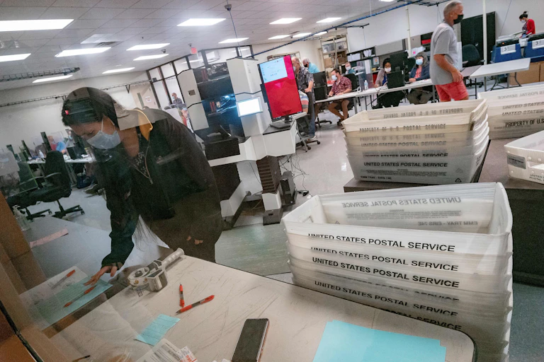 A woman in a face mask stands by piles of USPS boxes and looks at a ballot