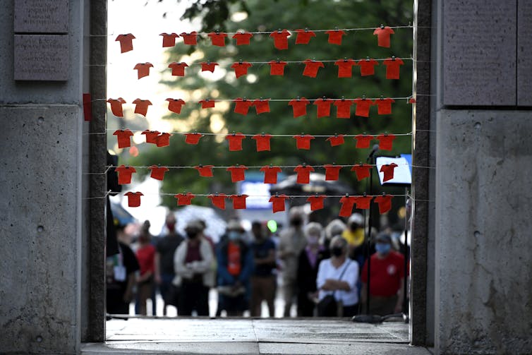 Orange fabric cut in the shape of shirts are pinned to string at the Human Rights Monument in Ottawa. They're strung across like clothes on a clothes line as people stand blurred in the background.