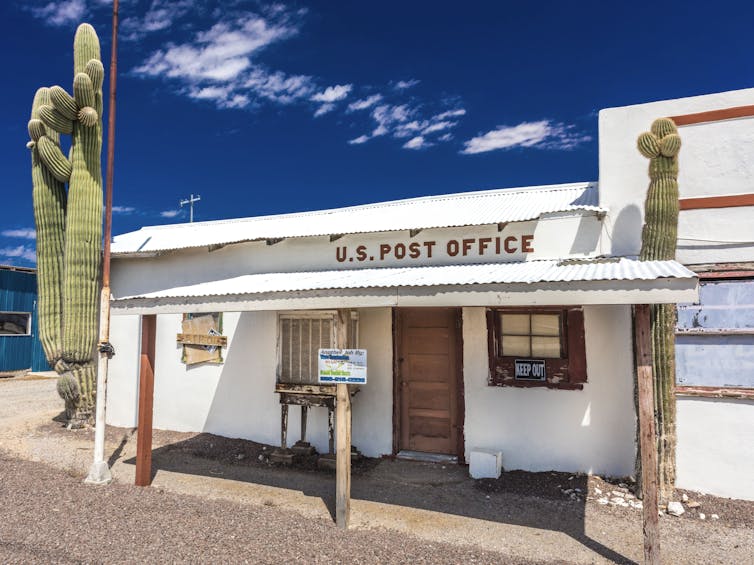 A small white post office that seems to be boarded up; a cactus is in front of it