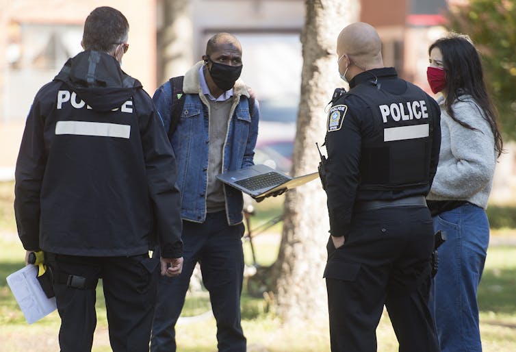 A man holding a laptop and a woman, both wearing masks, talk with two police officers.