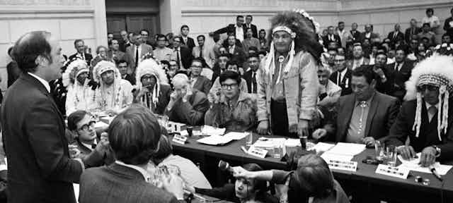 A black-and-white photo shows Pierre Trudeau standing and talking to Indigenous leaders.