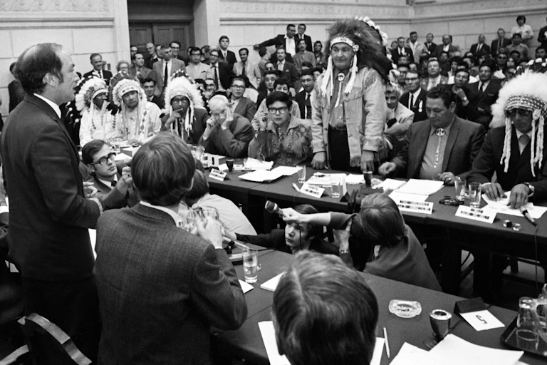 A black-and-white photo shows Pierre Trudeau standing and talking to Indigenous leaders.