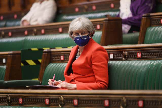Former prime minister Theresa May sitting on the green benches of the House of Commons wearing a COVID facemask with her glasses pull down over it.
