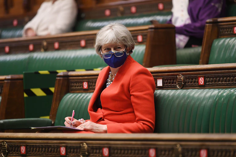 Former prime minister Theresa May sitting on the green benches of the House of Commons wearing a COVID facemask with her glasses pull down over it.
