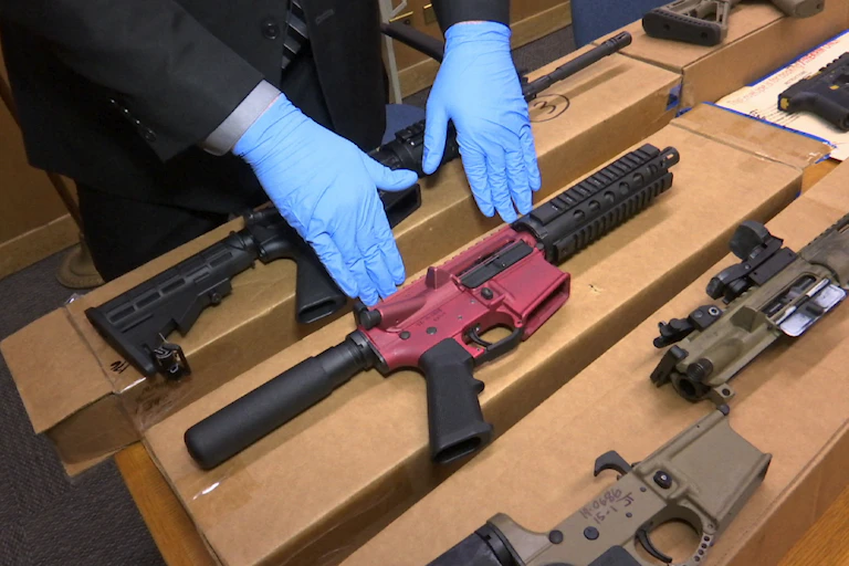 A man with gloved hands gestures at three firearms