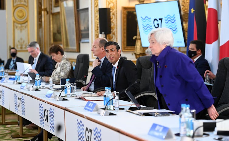 Ministros de Finanzas del G7 sentados en un banco largo con computadoras y micrófonos.