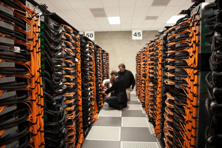 Rows of black boxes linked by orange cables flank a corridor where two people inspect a box