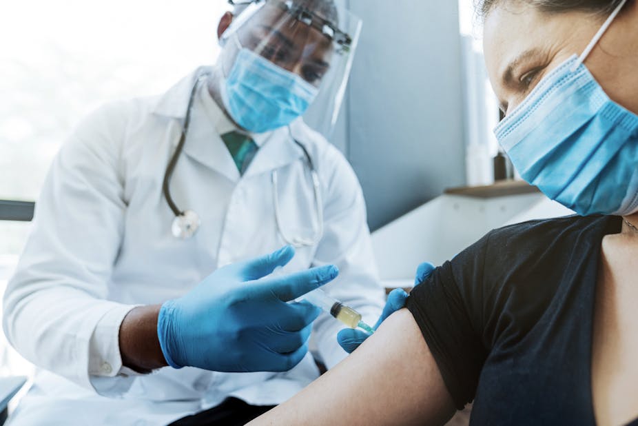 A doctor, wearing a mask and face shield, injecting vaccine on female patient's arm who's also wearing a face mask.