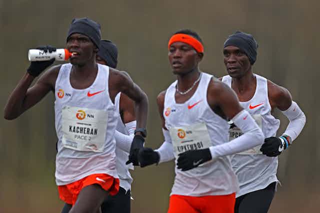 Four men running in white vests in a pack, an older runner at the back of the pack and at the front, younger runners in the middle.