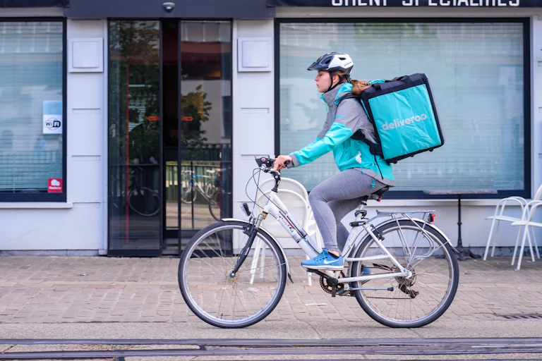 A woman on a bicycle with a large, square backpack with the Deliveroo logo.