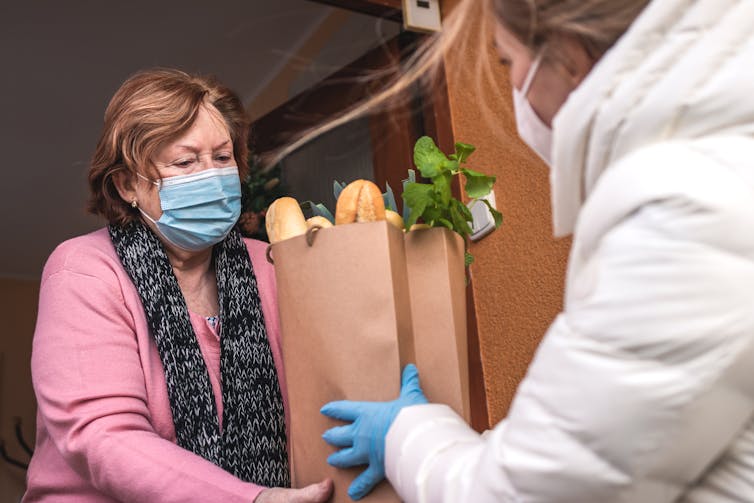 A woman hands a bag of groceries to another woman, both wearing face masks