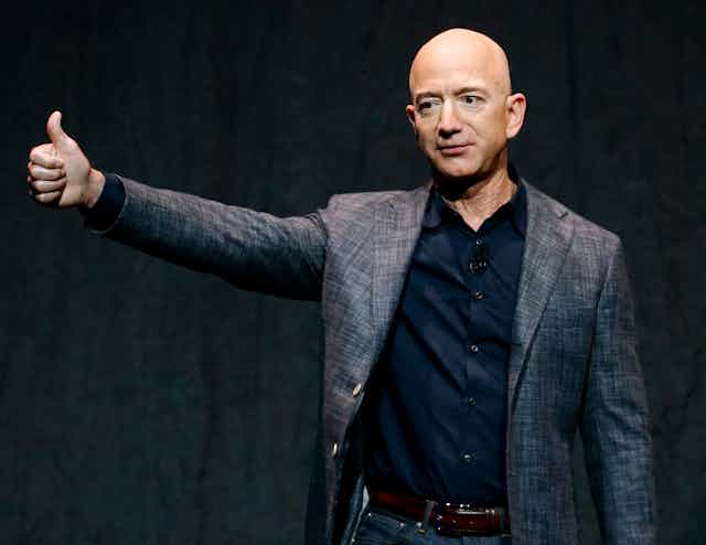 Jeff Bezos wearing gray sport jacket over a navy dress shirt, with no tie, holds his right arm up in the air in a thumbs up position