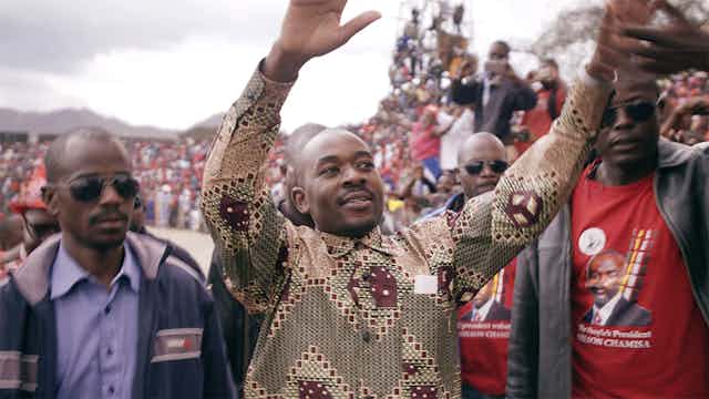 three men in front of a huge crowd. The man in the centre holds his hands up with a small smile. He is shaven headed and wears an African print shirt.