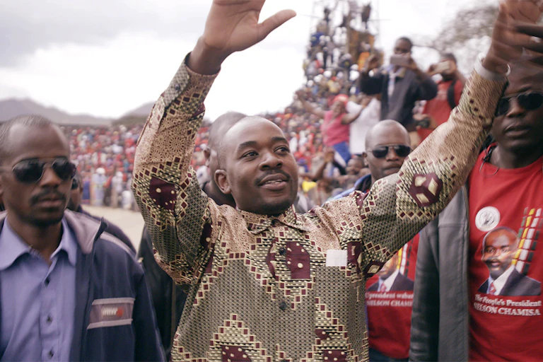three men in front of a huge crowd. The man in the centre holds his hands up with a small smile. He is shaven headed and wears an African print shirt.