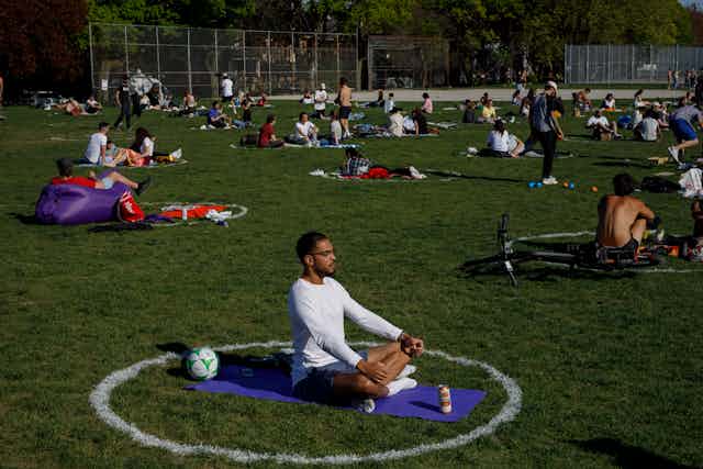 People sitting in white circles in a park. In the foreground a black man sitting on a yoga mat.