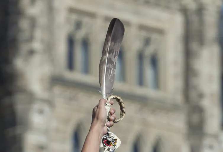 Woman holds up an eagle feather and sweetgrass braid in front of a bricked building that is blurred in the background.