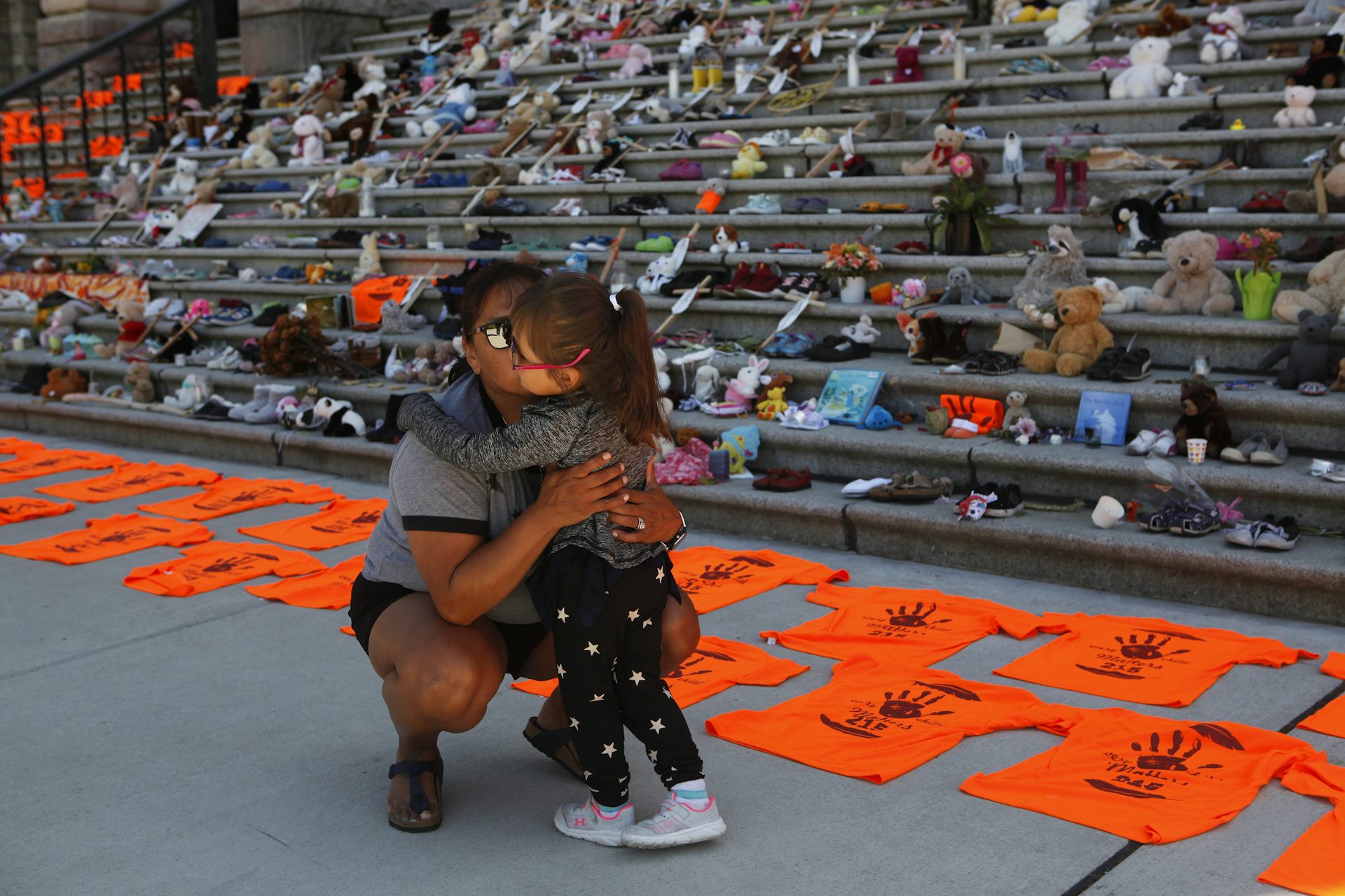Woman and daughter hug in front of steps covered in shoes and orange t-shirts