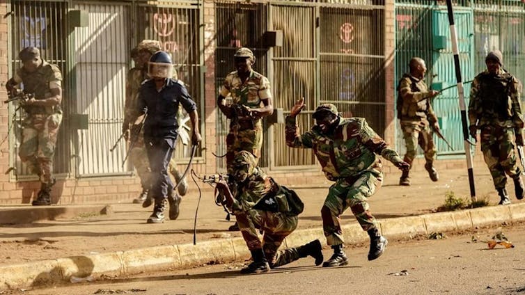A chaotic street scene featuring half eight soldiers in camouflage and a policeman. One soldier has kneeled and is shooting his rifle. Behind him another has his hand up to slap him on the back, laughing.