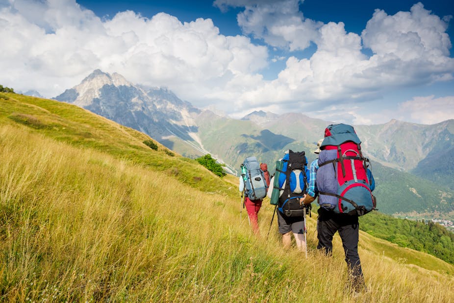 A group of three hikers with large backpacks climbs up a grassy hill.
