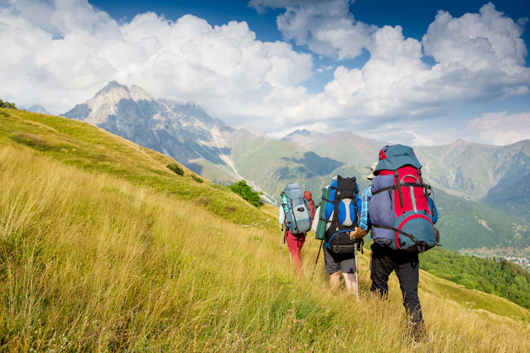 A group of three hikers with large backpacks climbs up a grassy hill.