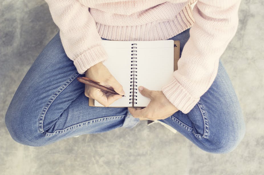 Girl wearing a cream jumper and blue jeans seen from the arms down sitting cross=legged on a concrete floor and writing in a notebook.
