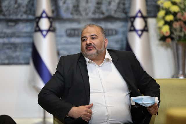Mansour Abbas, a middle-aged man with a beard, white shirt and black jacket, seated in front of two Israeli flags