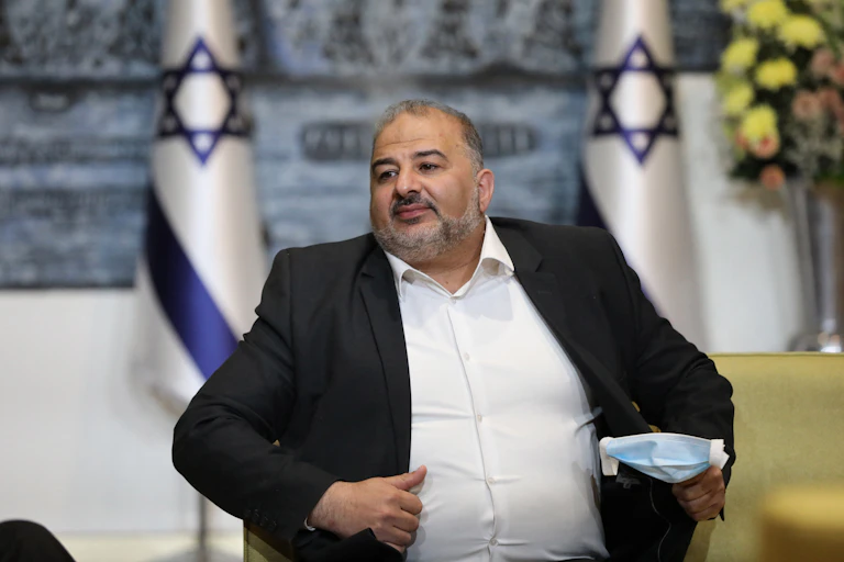 Mansour Abbas, a middle-aged man with a beard, white shirt and black jacket, seated in front of two Israeli flags
