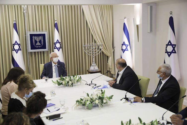 A group of people sit at a large table with white tablecloth and Israeli flags in the background.