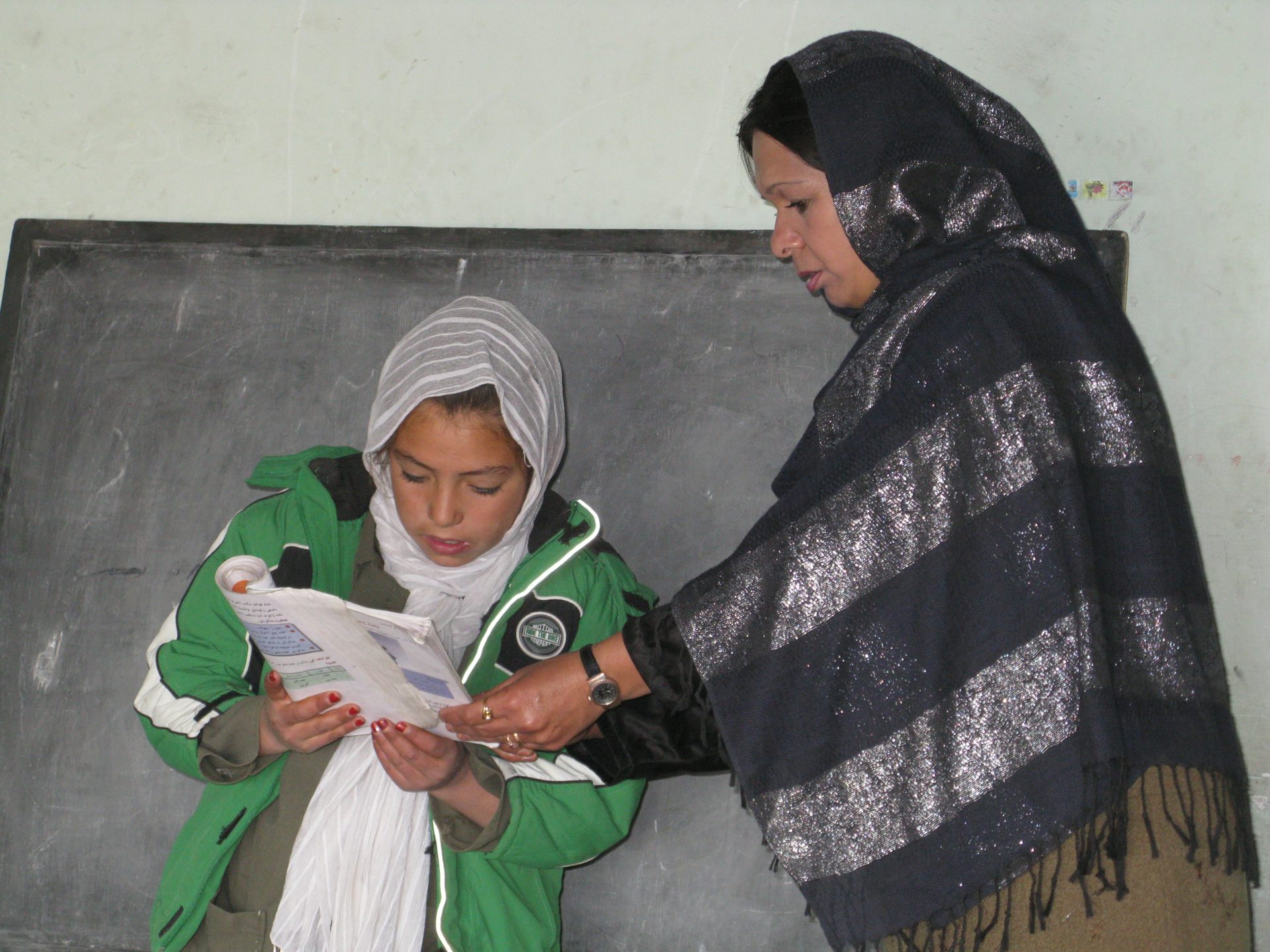 In front of a black board, a female teacher stands beside a girl and helps her hold a book, while the girl is reading. 
