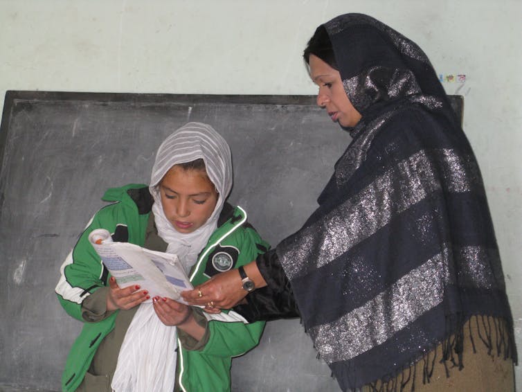 In front of a black board, a female teacher stands beside a girl and helps her hold a book, while the girl is reading. 