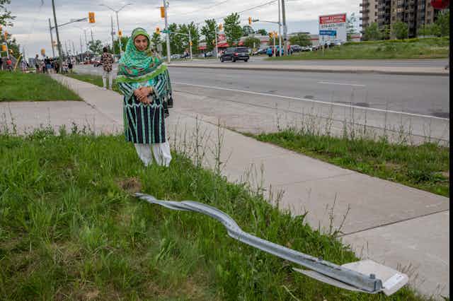 A woman wearing a hijab stands next to a destroyed street sign.