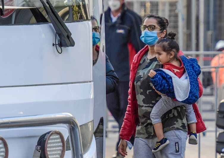 Women in mask carries a child on her hip as she boards a bus