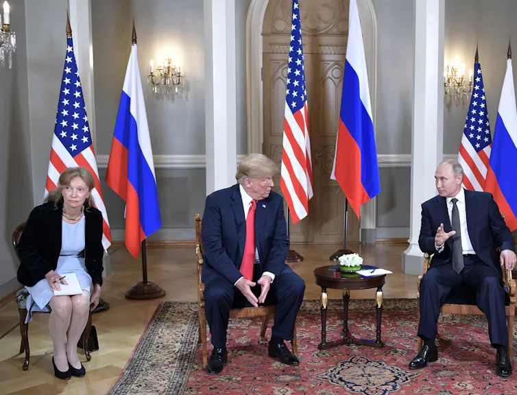 Donald Trump and Vladimir Putin sitting in chairs, in conversation. A US translator is sitting next to Trump, and American and Russian flags are in the background.