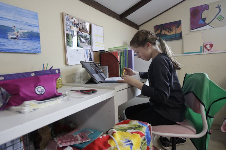 Un estudiante se sienta en un escritorio en el dormitorio de una casa, frente a una computadora portátil y con papel y lápiz en la mano