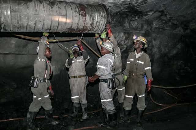 Men in helmets, overalls, boots and gloves stand in a tunnel underneath a large pipe