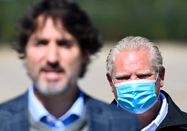Doug Ford, wearing a mask, is seen behind Justin Trudeau.