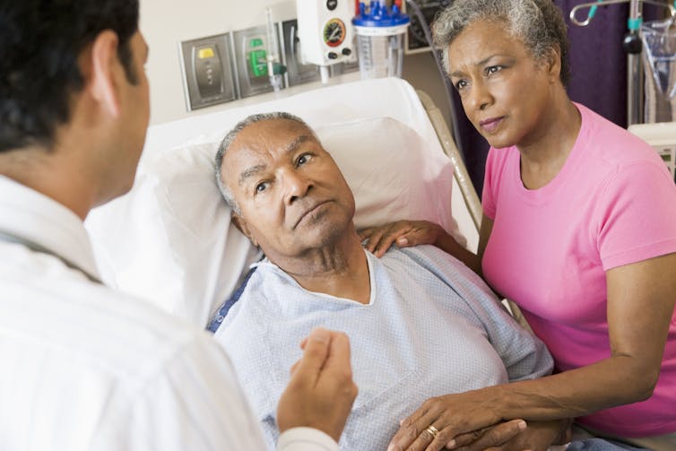 A man in a hospital bed and a woman holding his hand, listening to a doctor whose back is to the camera.