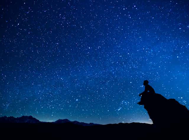 A person sits on a rock promontory against a vast night sky full of stars.