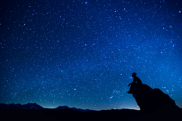 A person sits on a rock promontory against a vast night sky full of stars.