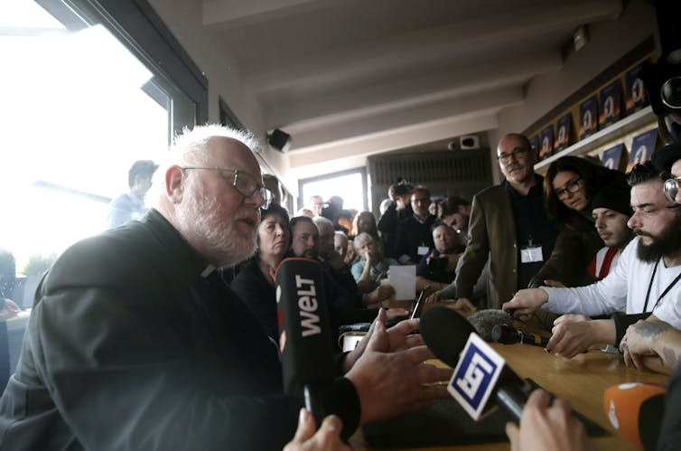 A man in a priest's collar and with a beard speaks with some reporters and a crowd.