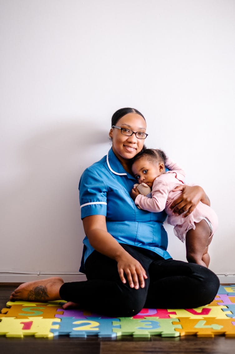 NHS midwife sitting on children's play mat while holding her daughter