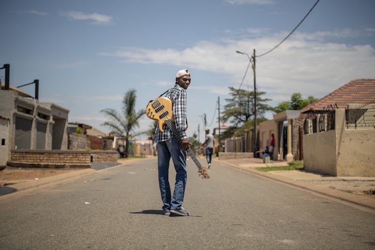 A township street and a blue sky; an elder man stands in the middle of the road and looks back to the camera, a guitar slung over his shoulder.